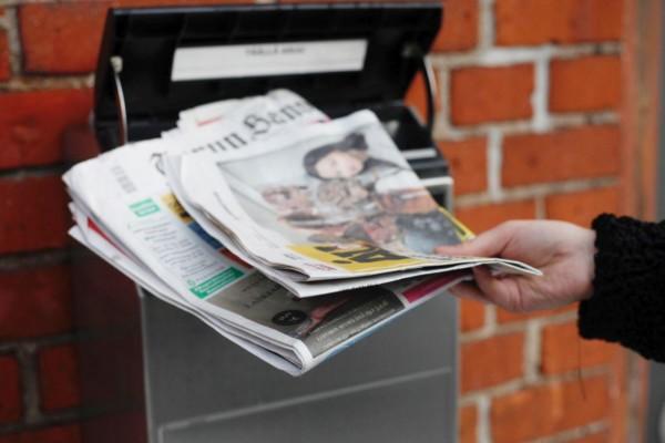 En hand med en bunt tidningar framför en postlåda.