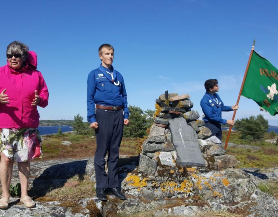 Kvinna och två ynglingar står vid monument på klippa