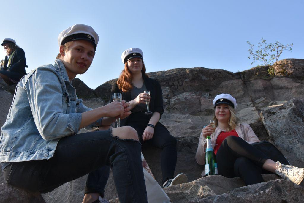 Tre ungdomar iklädda studentmössor sitter på en klippa och håller champagneglas i händerna.
