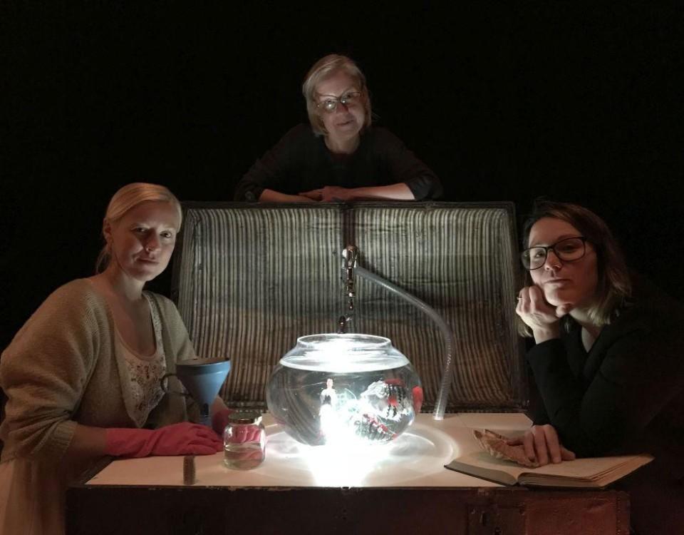 Tre personer står och sitter runt ett bord med en upplyst skål med vatten i mitten, svart bakgrund.