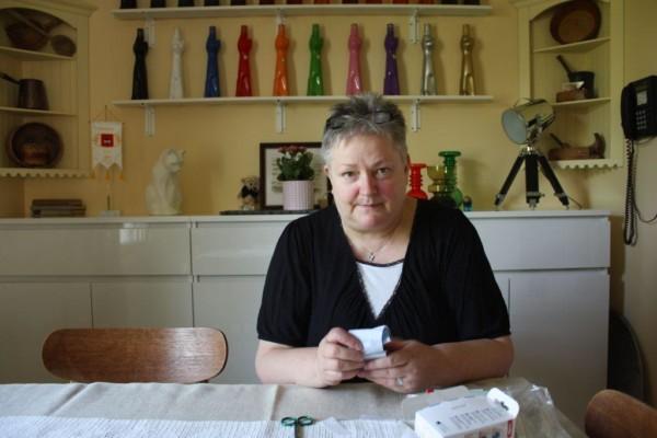Mona Söderblom sitter vid ett bord och håller en plsterförpackning i handen, i bakgrunden en vit byrå och inredningsprydnader
