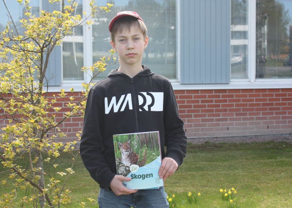 Samuel Engblom med röd keps på huvudet, håller i sin biologi bok med titeln "Skogen", i bakgrunden en buske och skolhuset.