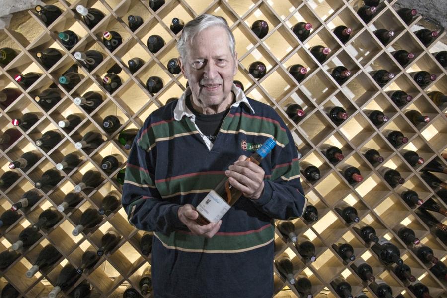 äldre man håller en vinflaska och bakom honom finns en större vinställning