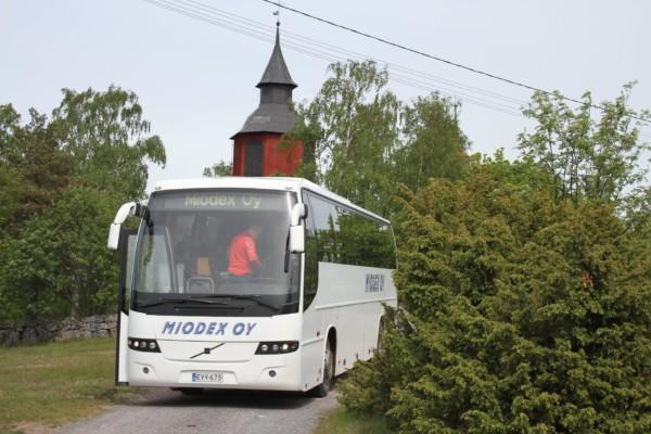 Framända på vit buss med texten Miodex, grönt buskage på sidorna, rött kyrktorn bakom bussen.