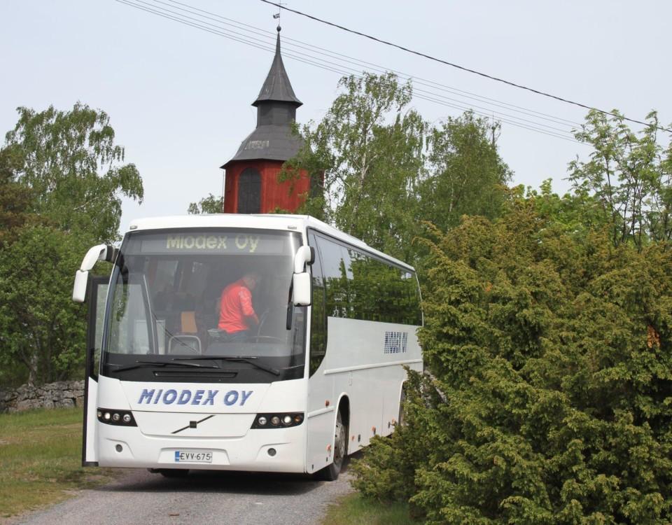 Framända på vit buss med texten Miodex, grönt buskage på sidorna, rött kyrktorn bakom bussen.