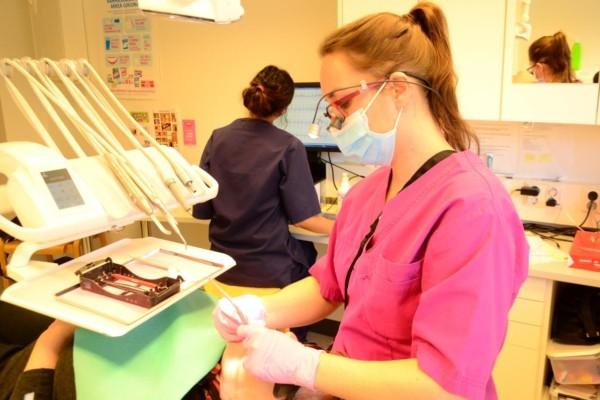 tandläkare undersöker en patients mun och i bild ses även en tandskötare som vid dataskärmen antecknar uppgifter