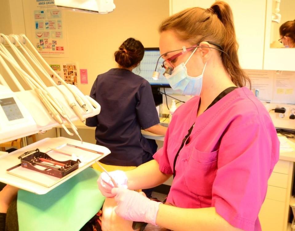 tandläkare undersöker en patients mun och i bild ses även en tandskötare som vid dataskärmen antecknar uppgifter