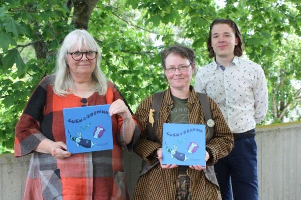 Tre personer står och ler in i kameran, Eivor Lindgård och Satu Zwerver med varsin blå barnbok i händerna, i bakgrunden ett grönt lövträd
