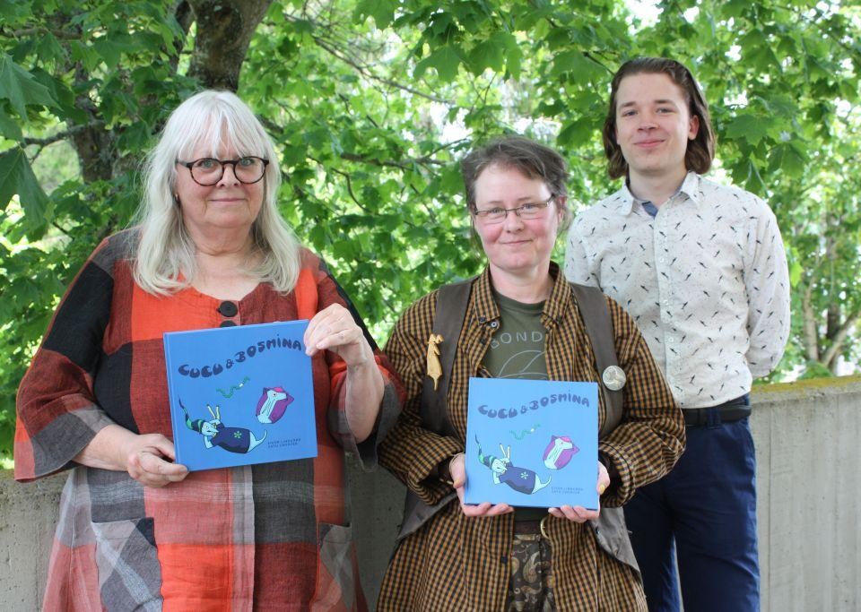 Tre personer står och ler in i kameran, Eivor Lindgård och Satu Zwerver med varsin blå barnbok i händerna, i bakgrunden ett grönt lövträd