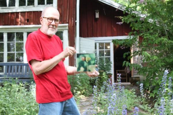 Janne Gröning håller i en glastavla med blomtryck, står utanför det röda huset i Westers Trädgård som han ställer ut i, grönt träd i bakgrunden och lila blommor runt omkring