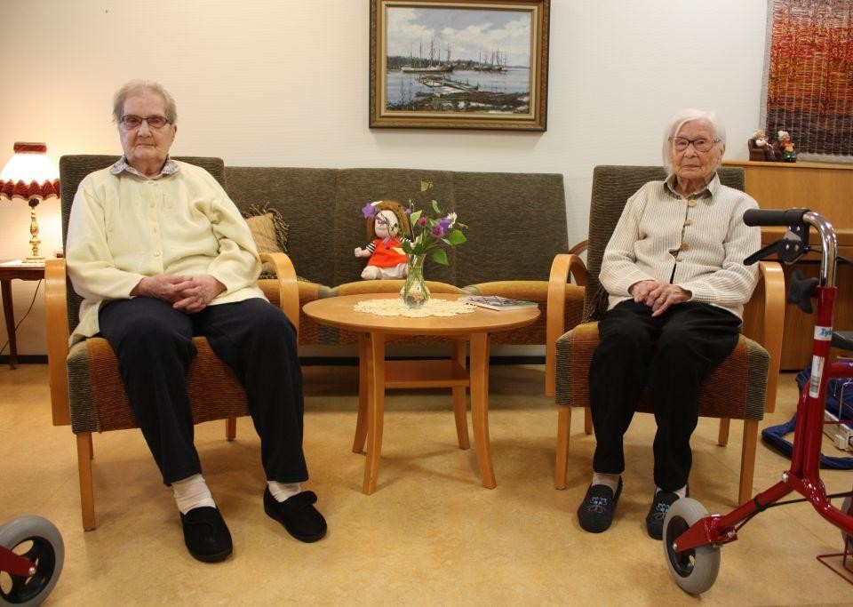 Margit Karlsson och Violet Åhlfors sitter i varsin stol, mellan dem står ett litet runt bord, i bakgrunden en grön soffa och en tavla på väggen