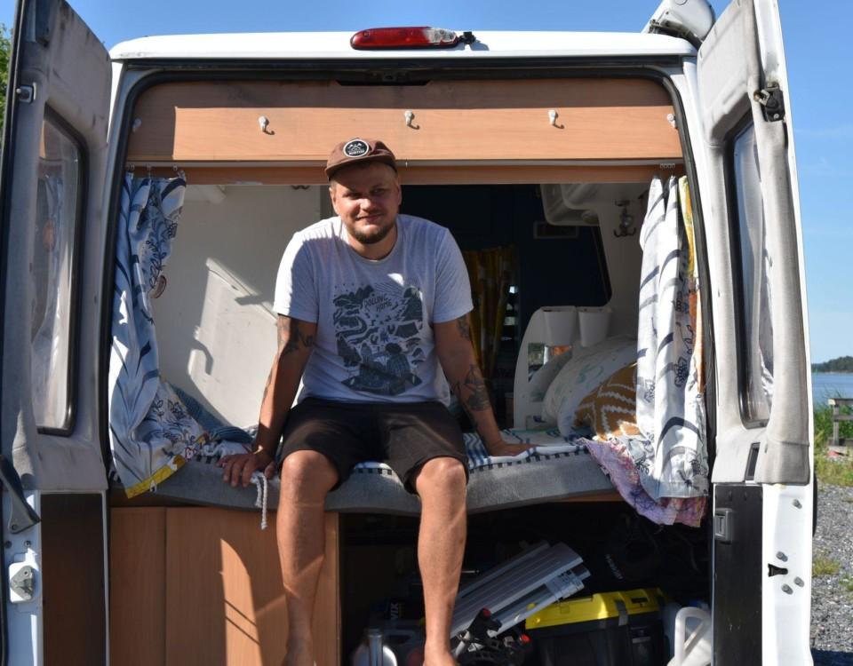 En man klädd i shorts, t-skjorta och keps sitter på en säng som finns i den öppna bakluckan av en paketbil.