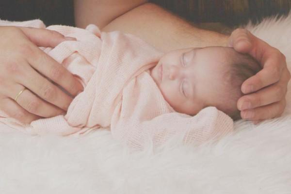 ett nyfött barn ligger inlindad i en filt och sover. Ett par vuxna händer håller om barnet.