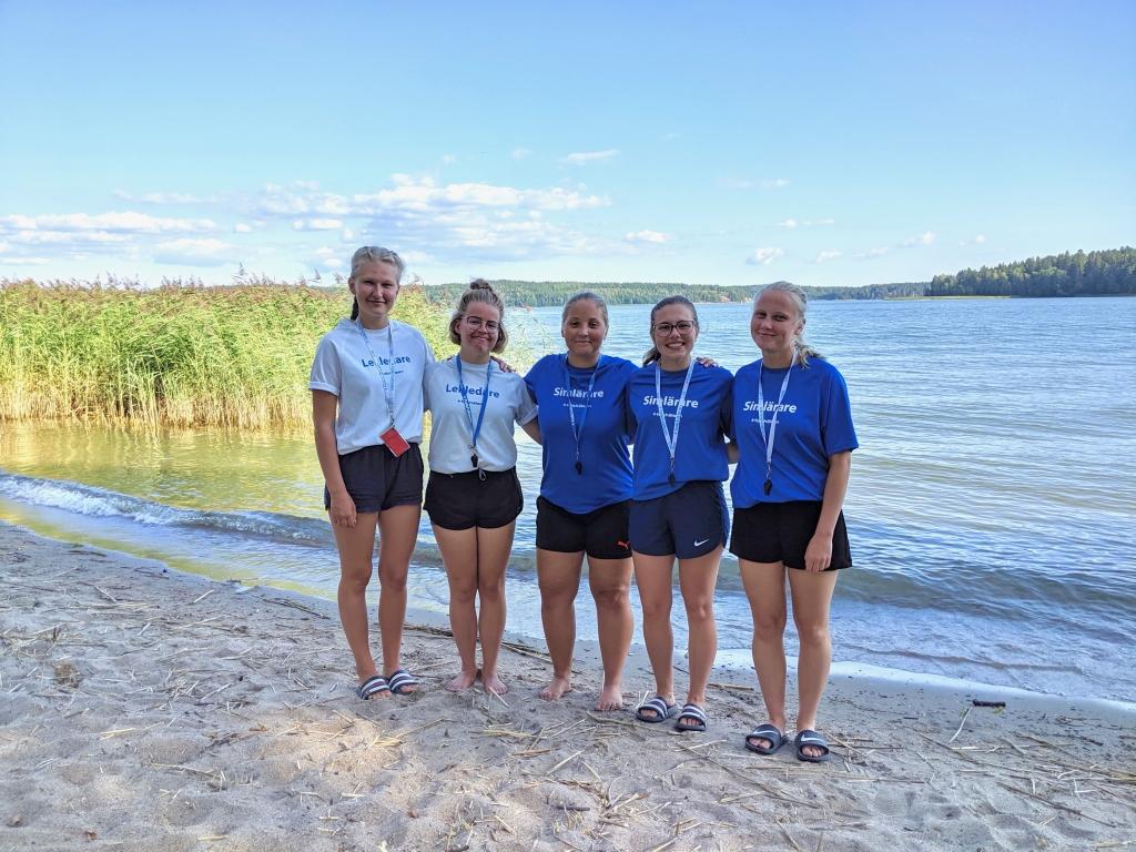 Fem unga kvinnor klädda i Folkhälsan-skjortor står och håller om varandra på en strand.