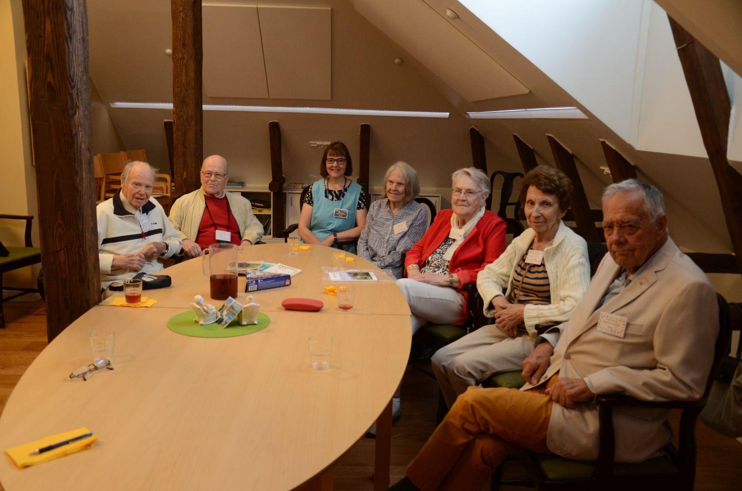 En grupp seniorer samt gruppledaren sitter kring ett bord och ler mot kameran.