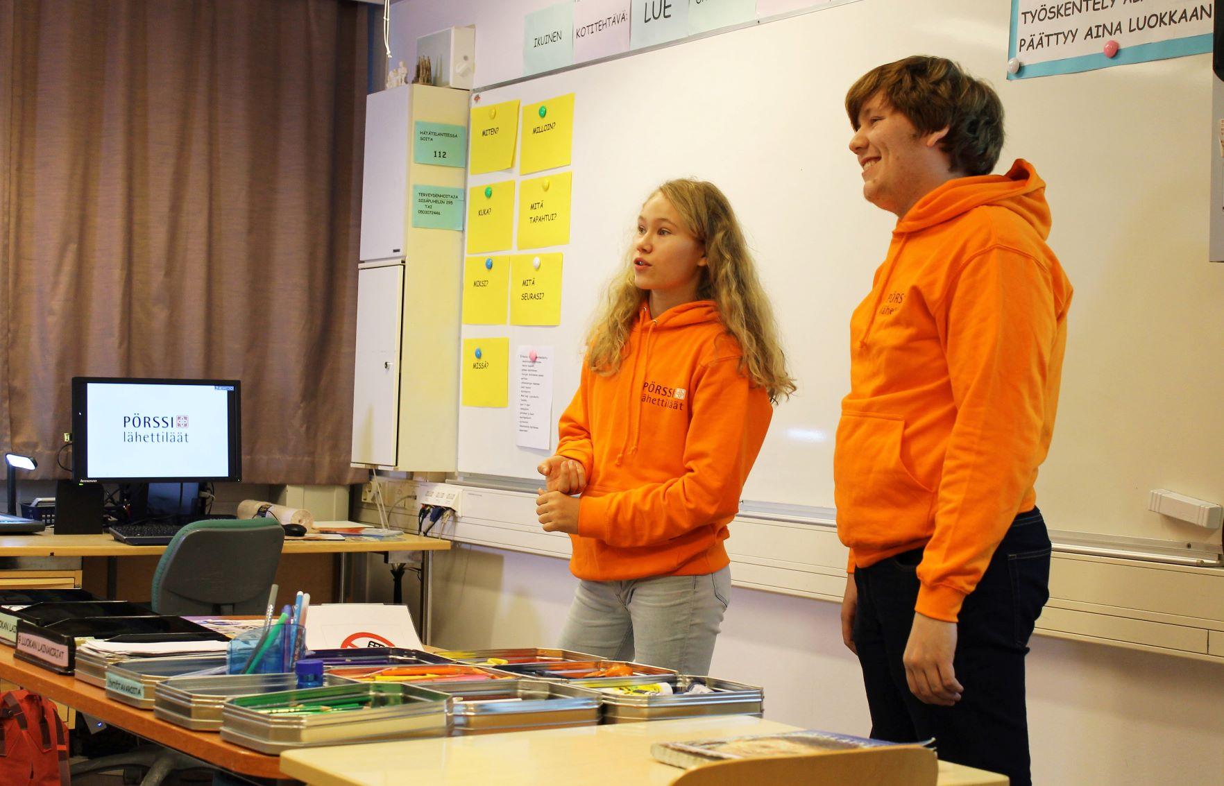 Två unga i orangea collegar står framme i klassen