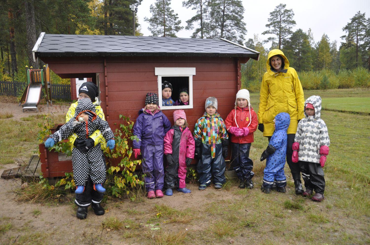 Ett gäng med 10 barn står vid en lekstuga i regnet tillsamman med en vuxen