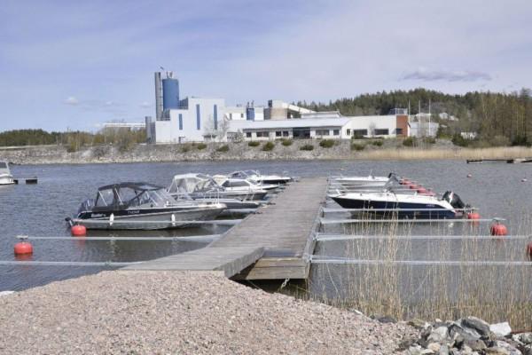 I bakgrunden syns fabriksbyggnad. Framför en vik och en brygga med båtar.