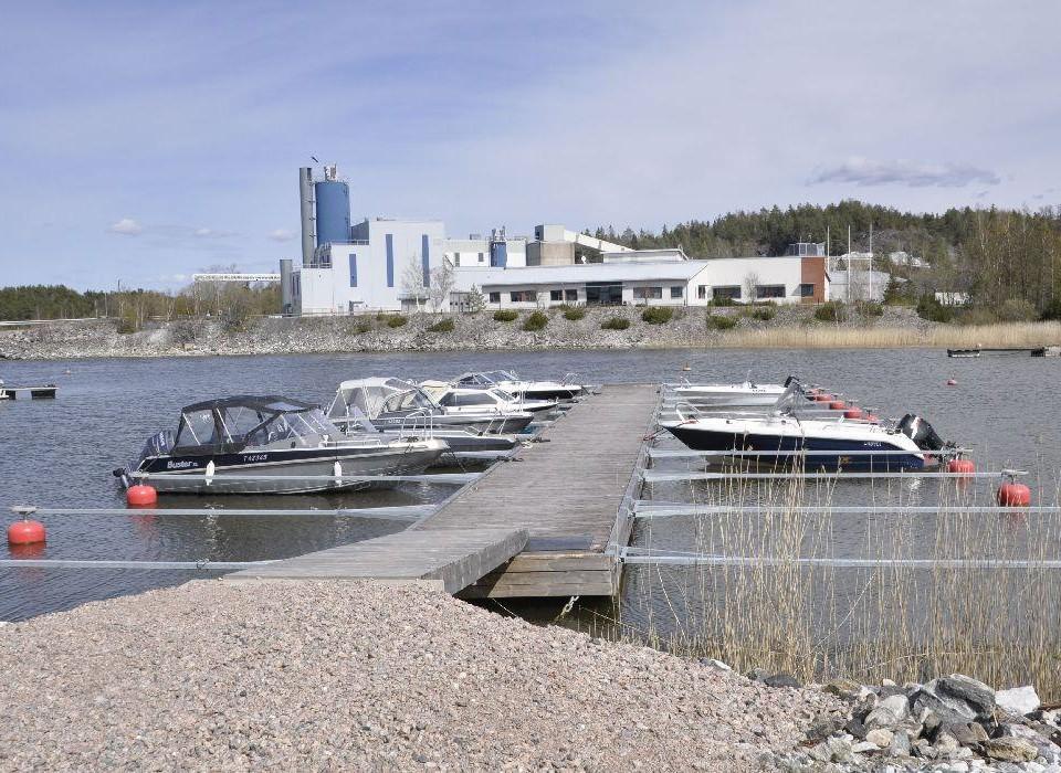 I bakgrunden syns fabriksbyggnad. Framför en vik och en brygga med båtar.