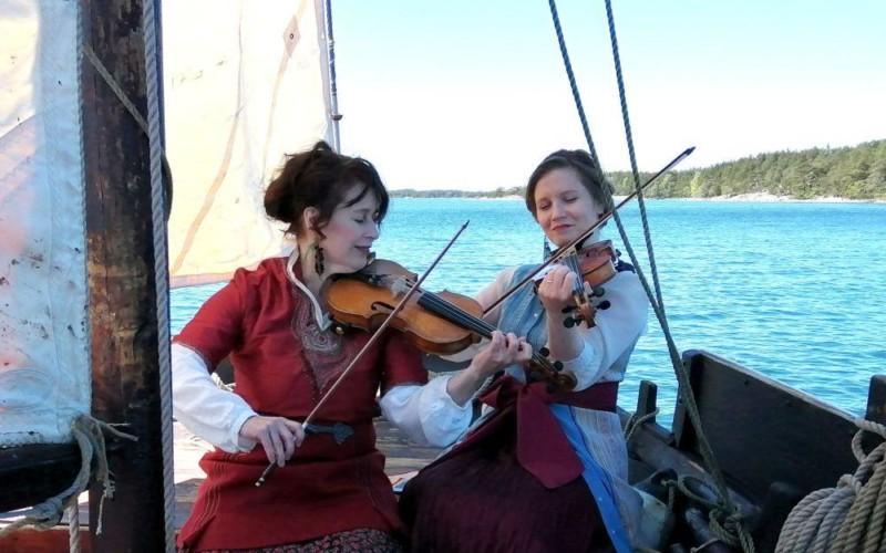 Två kvinnor i traditionella kläder sitter ombord en segelbåt och spelar fiol.
