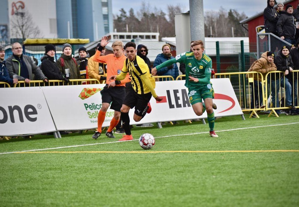 Två manliga fotbollsspelare, en i gul tröja och en i grön, jagar en boll på en fotbollsplan.