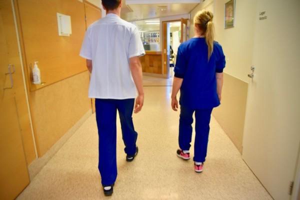 Ryggen av två personer klädda i vårdpersonalskläder i en sjukhuskorridor.