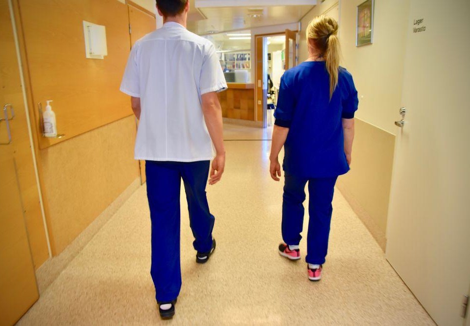 Ryggen av två personer klädda i vårdpersonalskläder i en sjukhuskorridor.