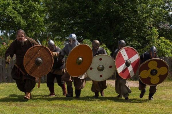 En grupp människor klädda i vikingakläder och bärandes sköldar springer över en gräsmatta.