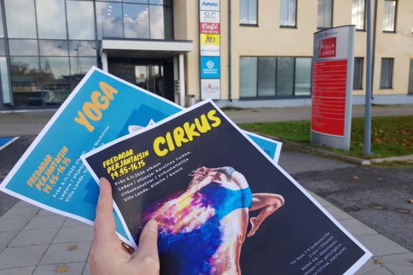 Två flyers- cirkus och yoga - i en hand framför ett kulturhus