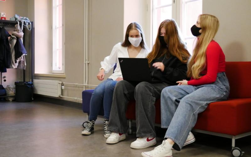 tre gymnasieelever med munskydd sitter samlade kring en laptop på en soffa