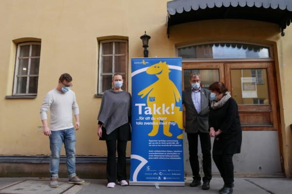 fyra personer med munskydd står samlade kring en stor banderoll på en gul filur och ordet "Takk" med två k