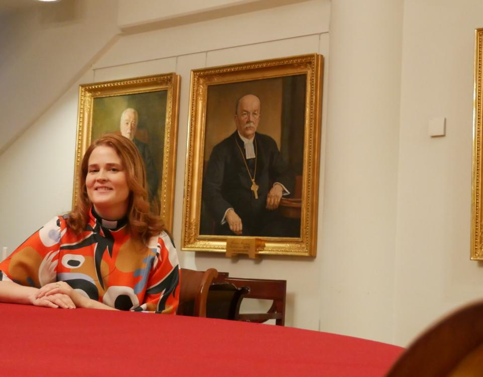 en kvinna sitter vid ett bord med en röd duk, i bakgrunden målade portrtätt av äldre, manliga präster