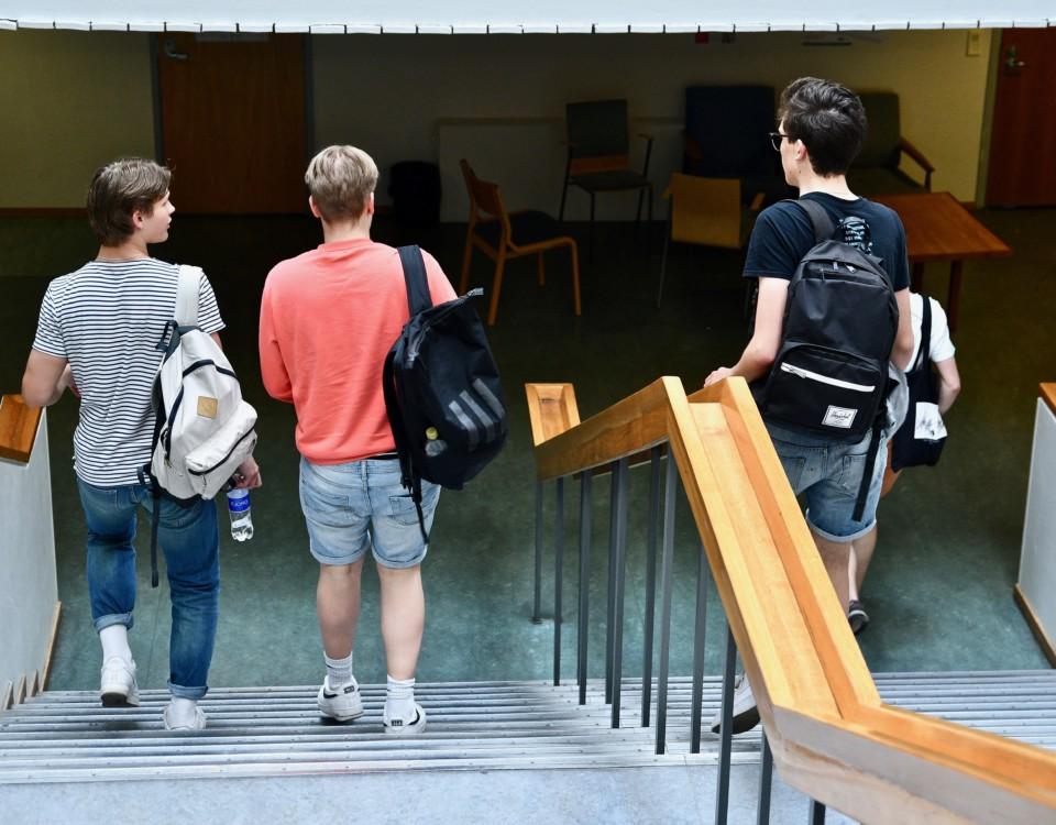 Tre unga killar med ryggsäck på ryggen går nerför en trappa i en skola.