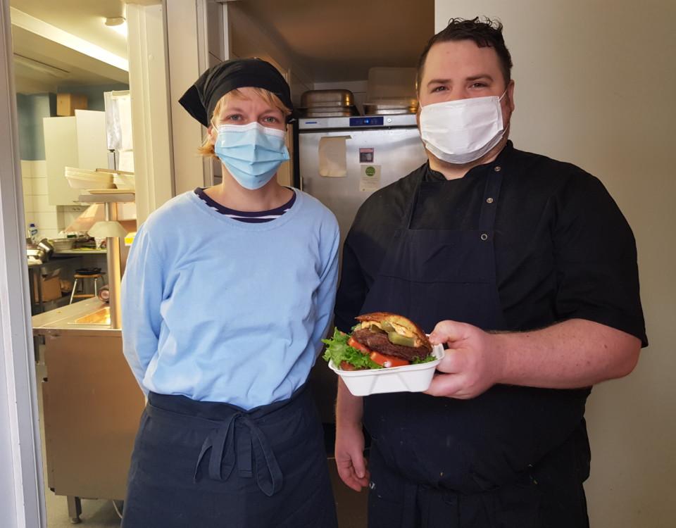 Två personer som jobbar i ett kök poserar med en hamburgare i en liten låda.