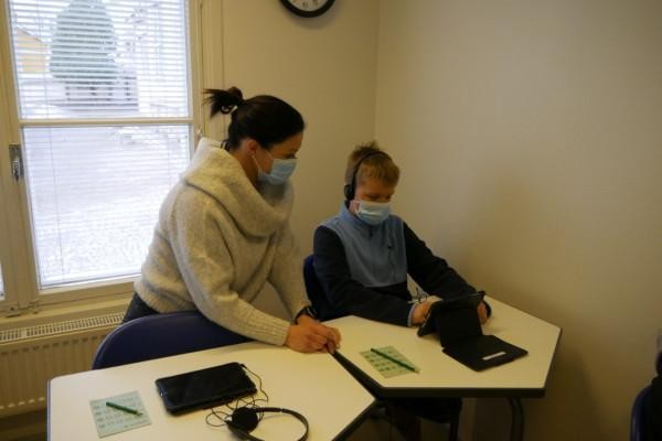En pojke sitter med hörlurar och tittar på en skärm medan en kvinna sitter bredvid. Båda bär munskydd.