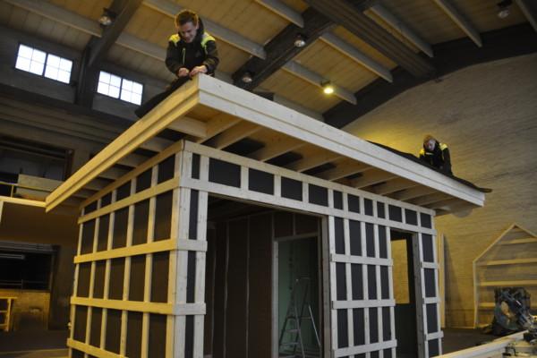 Ett modulhus under arbete i en hall. PÅ taket två unga arbetande män