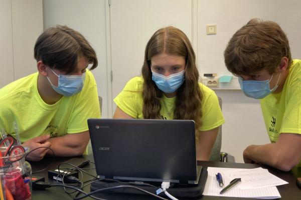 tre skolelever med munskydd framför en bärbar dator