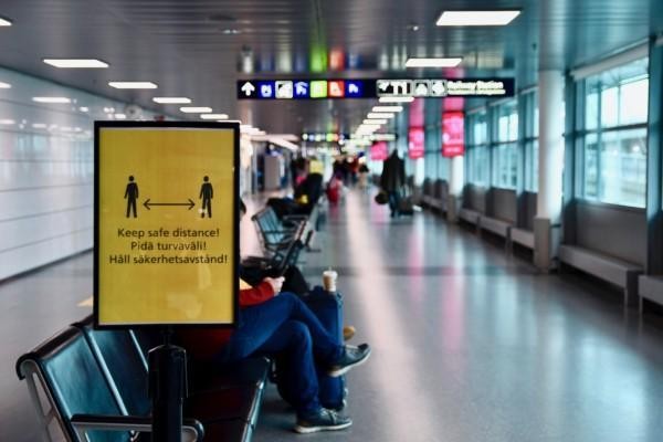 En vänthall på ett flygfält. En gul skylt påminner om att man bör hålla säkerhetsavstånd.