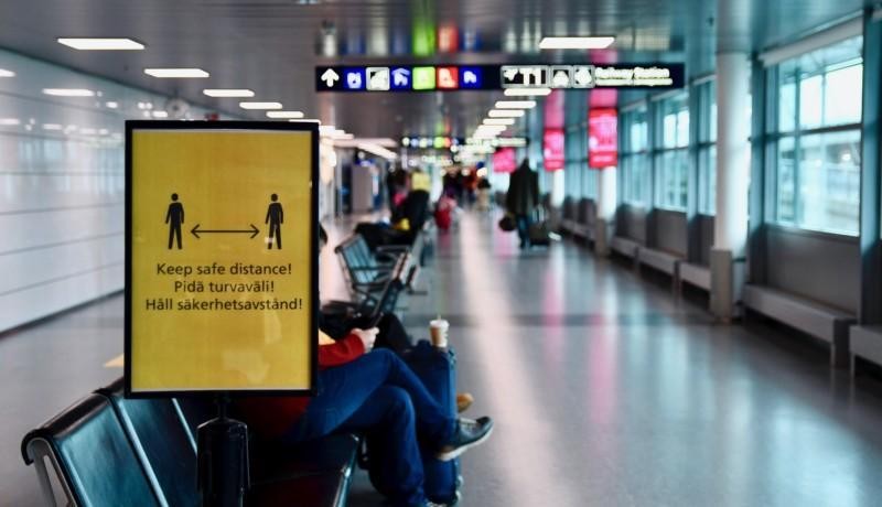 En vänthall på ett flygfält. En gul skylt påminner om att man bör hålla säkerhetsavstånd.