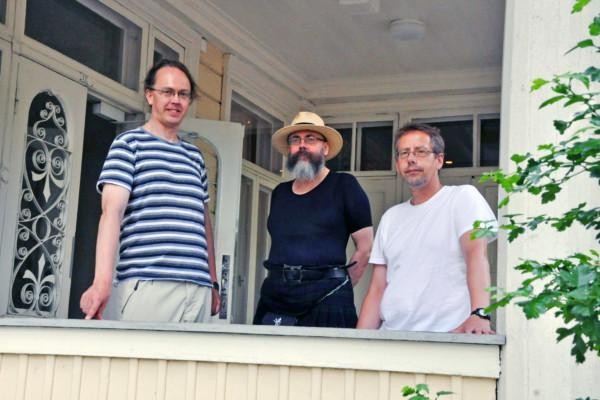 Tre män står på en terrass och tittar in i kameran.
