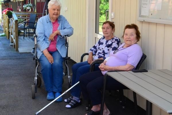 Tre kvinliga pensionärer sitter i skuggan mellan två hus. De tittar in i kameran