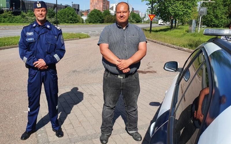 Peltola iklädd polisuniform och Heiskanen i skorta och jeans poserar framför en polisbil med motorvägen i bakgrunden.