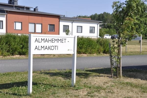 Skylt med texten "Almahemmet - Almakoti", rödvitthus i bakgrunden, grönt buskage runt omkring