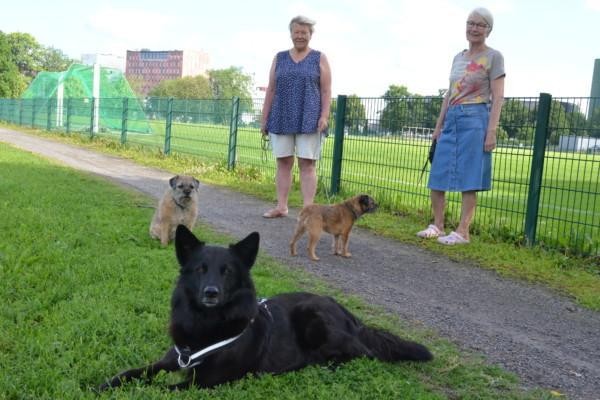 Två damer står på en gräsmatta med sina tre hundar, två små och en större svart