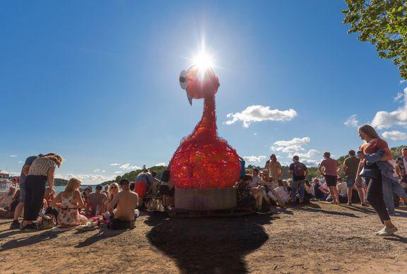 Ett stort konstverk i form av en flamingo står på en strand. Runt omkring sitter folk.