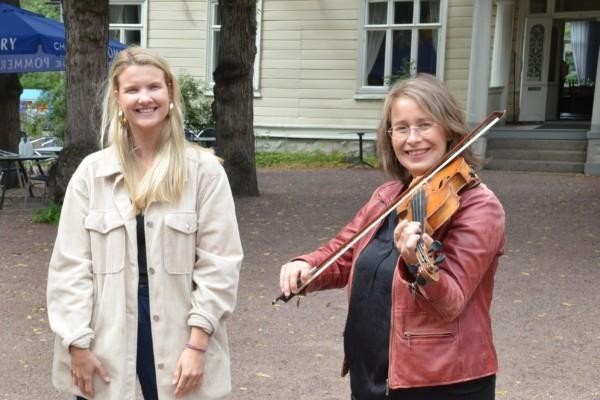 Två kvinnor står framför ett hus, en av dem spelar fiol