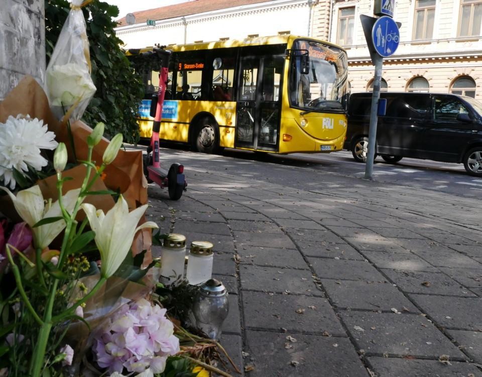 blommor och ljus på en trattoar, buss och parkerad elsparkcykel i bakgrunden