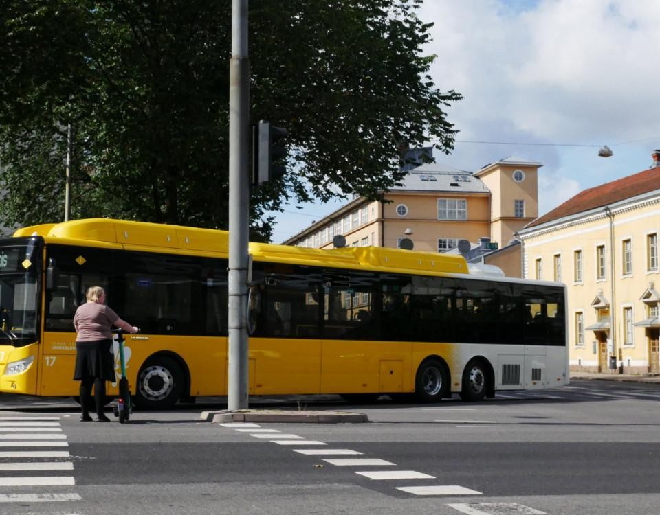 en buss gör en sväng och en person med elsparkcykel väntar på att gå över vägen
