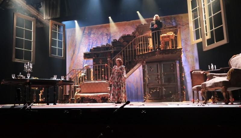 Bild av teaterscen. En kvinna i blommig klänning och en skäggig man på en trappa står på scennen.
