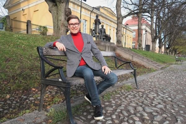 Bild av en man i bleiser och polotröja som sitter på en bänk vid åstranden i Åbo.